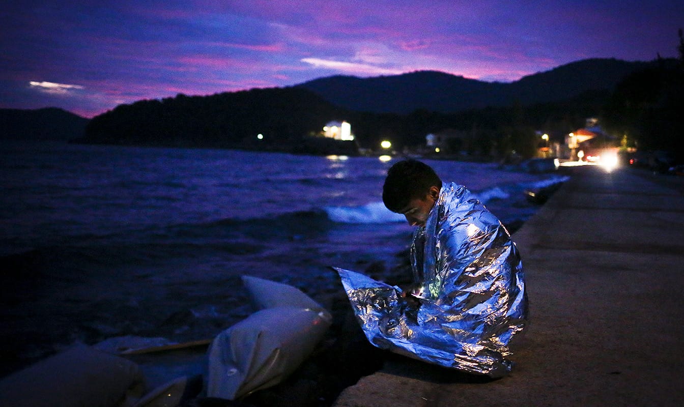 A Syrian refugee sits along a seashore at dusk, wrapped in a blanket.
