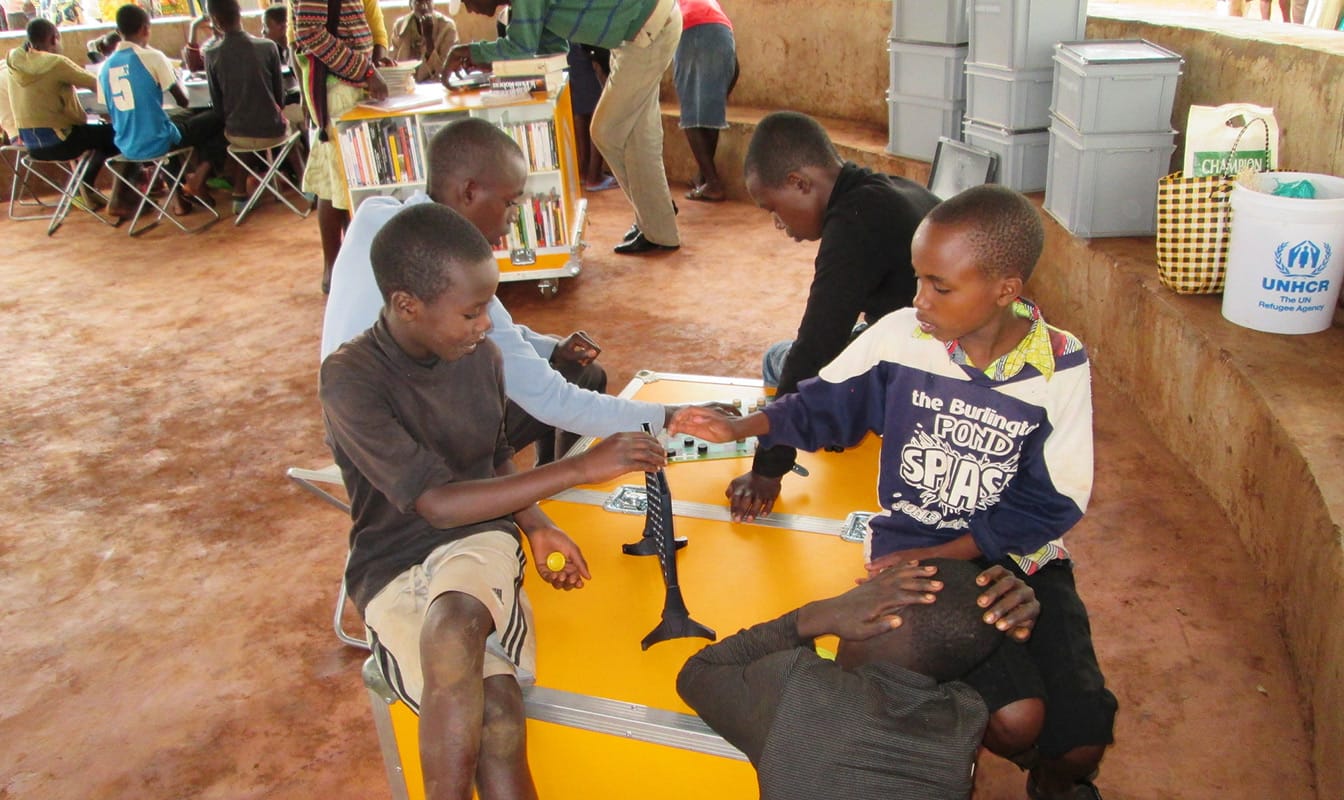 Five refugee children huddle around an Ideas Box, using its surface to play board games.