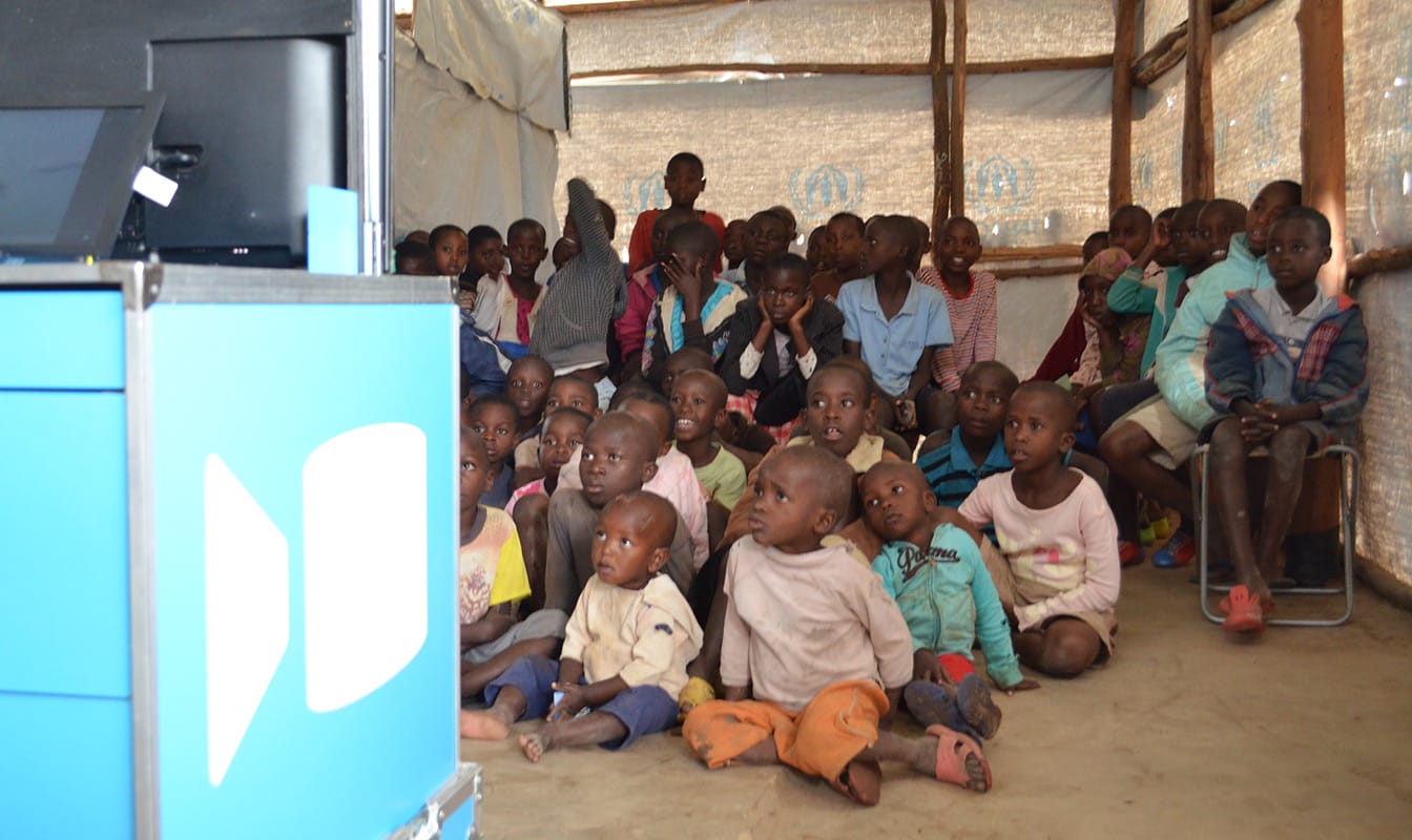 A large group of refugee children watches a television that is placed on top of an Ideas Box.