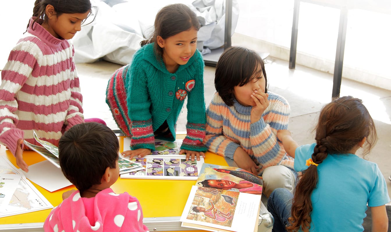 Five refugee children huddle around an Ideas Box reading picture books.