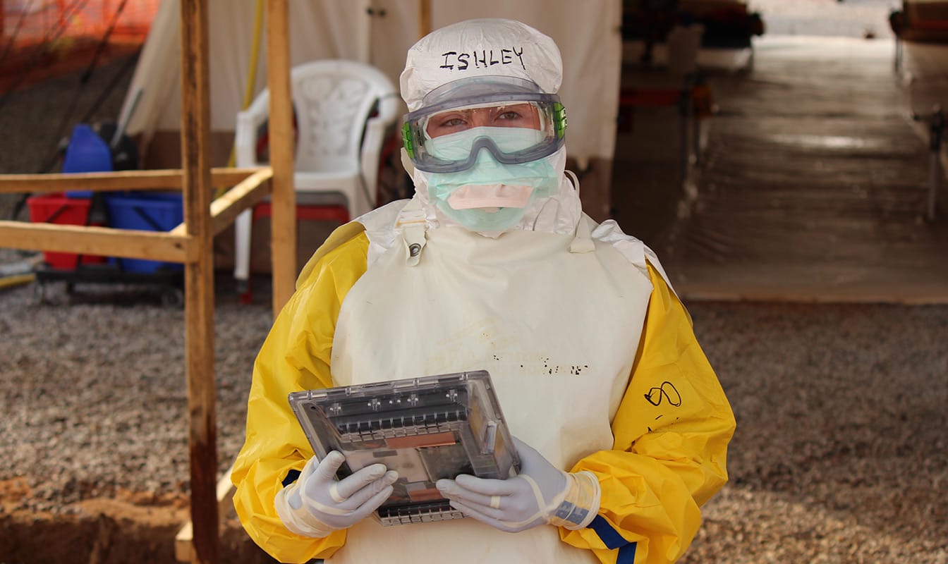 An MSF field worker in protective gear looks into the camera while using a tablet to access Project Buendia.